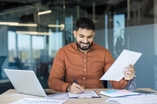 Young smiling businessman at his modern office desk signing documents, surrounded by laptop, charts and paperwork as he reviews financial data and plans strategy