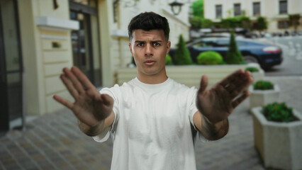 Man standing on city street with hands raised as if signaling stop wearing a white shirt in an...