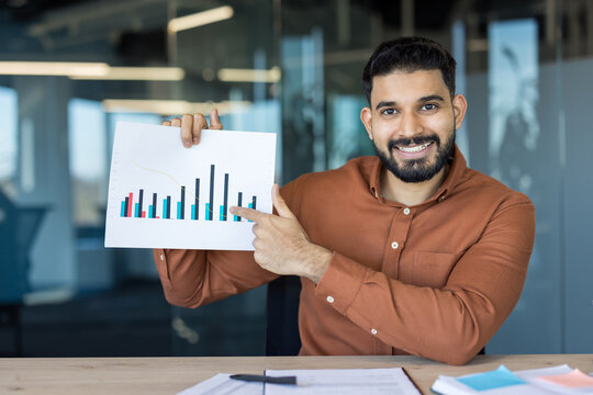 Smiling young indian businessman holding up a graph and pointing, explaining financial statistics during a presentation in a modern office setup, analyzing market trends