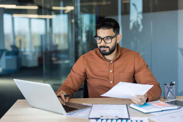 Fototapeta premium Businessman sitting at a desk in a modern office, working on a laptop while holding and reviewing paper documents, demonstrating dedication and productivity