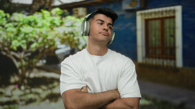 Young man wearing headphones in casual white shirt stands thoughtfully outdoors in urban street setting with blurred greenery and blue building background.