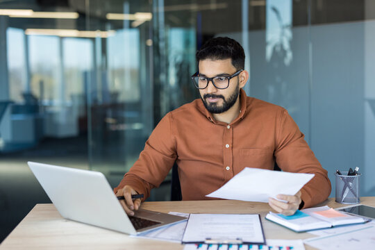 Businessman sitting at a desk in a modern office, working on a laptop while holding and reviewing paper documents, demonstrating dedication and productivity - Powered by Adobe