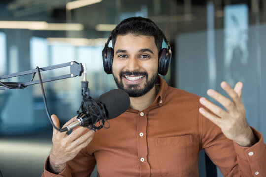 Young man wearing headphones and a rust-colored shirt, smiling and gesturing while talking into a professional microphone, actively creating audio content in a modern studio environment