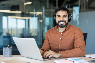 Obraz premium Young indian man in a modern office wearing headphones, smiling at camera while typing on a laptop, engaging in remote work, online learning, or virtual communication