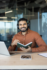 Indian man working on a laptop at a desk, smiling with headphones, holding a notebook, and engaging in online education or professional development in a modern office environment