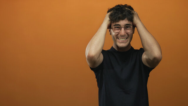 Young man wearing glasses and a black shirt clutching hair with both hands in studio orange wall; frustration.