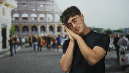 Young man with hands pressed to cheek for sleep, wearing glasses and black tshirt, posed in front of coliseum building amid tourists and blurred crowd; serenity.