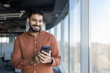 Indian man smiling and interacting with his mobile phone, standing in a modern office space with large windows, representing business communication and digital technology