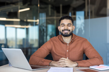 Young indian businessman seated at his desk with eyes closed and serene smile, practicing mindfulness and stress relief for calm, focus and well-being during a busy workday