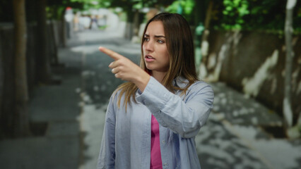 Woman pointing outdoors on a tree-lined street, wearing a casual shirt and looking serious in a urban setting, capturing a moment of focus and direction.