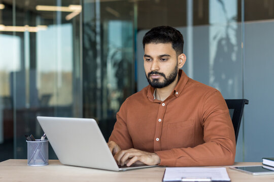 Young indian man staying focused while typing on a laptop at his desk, demonstrating dedication and productivity in a contemporary corporate environment