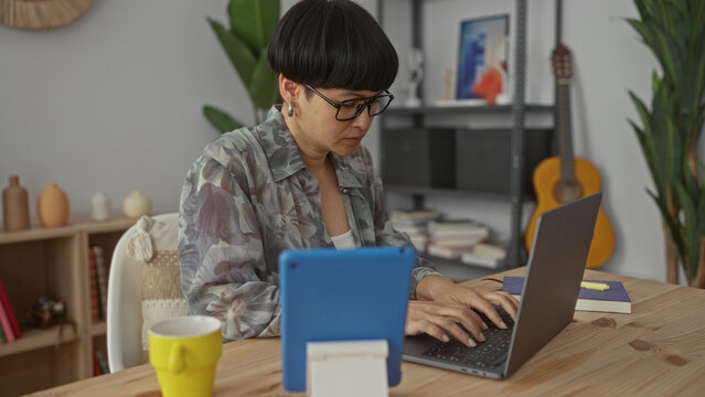 Woman working on laptop at home with tablet on desk in cozy living room setting including books and guitar implying a peaceful indoor environment.