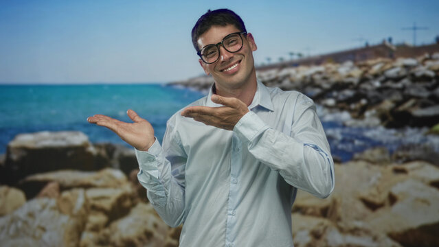 Man wearing glasses in white shirt smiles and points open palms at rocky shore and blue sea in studio; optimism. - Powered by Adobe