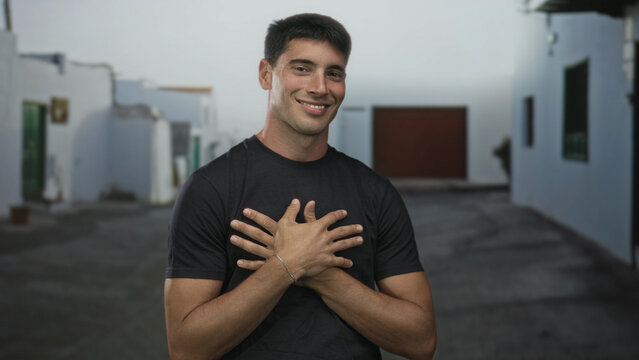 Smiling hispanic man wearing black shirt holds hands to chest on historic stone street in old town; self love gratitude. - Powered by Adobe