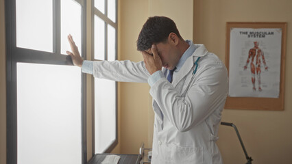 Fototapeta premium Man with stethoscope and white coat leans on window frame pressing palm to forehead in clinic; fatigue.