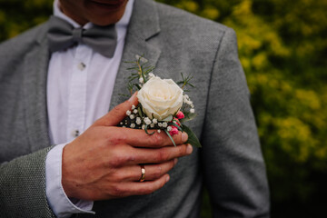 Man in gray suit with bow tie gently holding a white rose boutonniere adorned with greenery,...