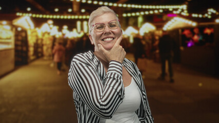 Middle aged woman smiling with hand on chin at night market street amid souvenir stalls and string lights; joy thinking.