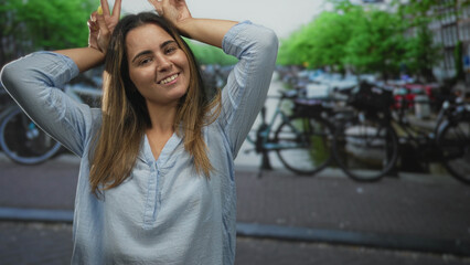 Fototapeta premium Young woman smiling with face and hands making bunny ears on amsterdam street by parked bicycles; playful carefree.