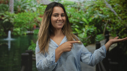 Obraz premium Woman smiling with palm up hand and pointing finger on sunlit forest walkway near pond in green park; joyful welcome.