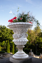 A decorative stone vase with red flowers and greenery standing on a terrace in Valea Morilor Park, Chisinau, Moldova. The ornate classical design, soft natural light, and lush green background create 