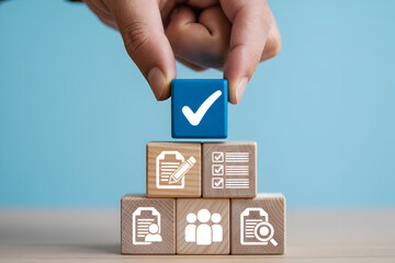 Hand places a blue block with a checkmark on top of wooden blocks with icons representing tasks teamwork and document review against a light blue background symbolizing success and completion