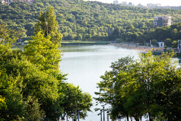 A scenic view of a lake surrounded by lush green hills, photographed from an elevated park area in Moldova. The peaceful landscape includes trees, distant buildings, and a sunny lakeside shoreline.