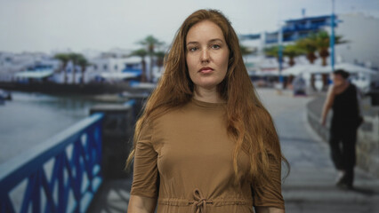 Young caucasian woman with hand on chin on seaside street under warm sunlight looking into distance; contemplation.