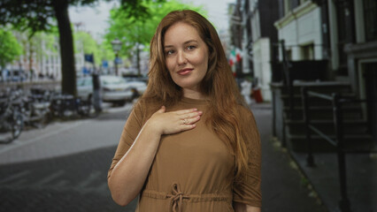 Smiling woman places hand on chest wearing brown dress on quiet city street in early morning light; gratitude.