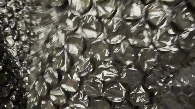 A hand crumples black plastic bubble wrap on a wooden table, creating textured movement and tactile sound in a close-up horizontal shot.