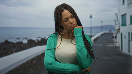 Woman with hand on chin and folded arms on seaside street promenade with ocean view; contemplation solitude calm.