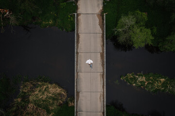 Fototapeta premium Aerial view of a solitary figure walking on a concrete bridge surrounded by lush greenery and dark water, capturing a serene moment in nature's embrace