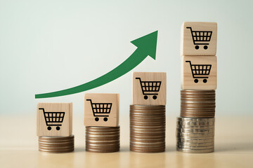 Rising sales represented by stacks of coins and shopping cart icons on wooden blocks with a green arrow indicating growth in ecommerce and retail business performance and financial success