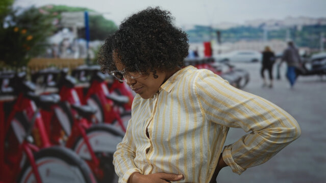 Young african american woman grips lower back beside rows of red rental bikes at bike parking on street; discomfort. - Powered by Adobe