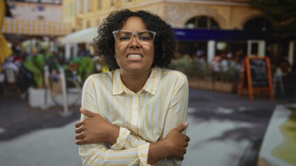 Woman wearing yellow striped shirt hugging herself with tense expression on restaurant terrace outdoors; discomfort.