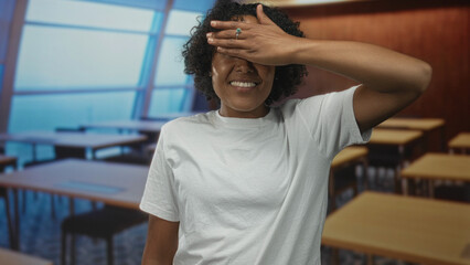 Woman in white shirt covers eyes with hand amid empty desks in building under soft sunlight; introspection calm.