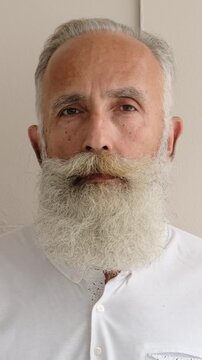Extreme close-up portrait of a serious senior man with a large white beard looking up and directly at the camera. Vertical video.