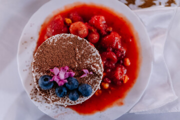 Delicious dessert featuring a creamy cake topped with fresh blueberries and edible flowers, served with a vibrant strawberry sauce on a white plate, showcasing culinary artistry and indulgence