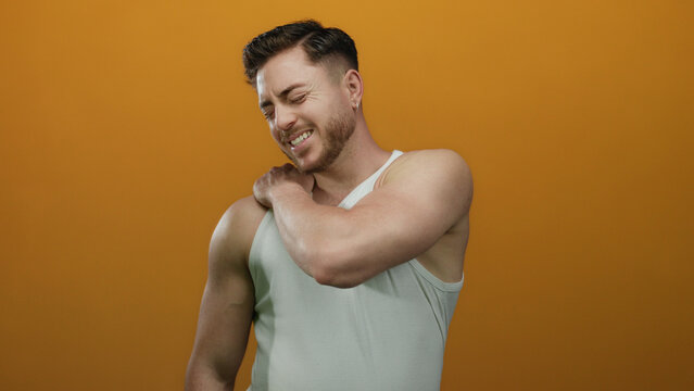 Young man with beard in white tank top experiencing shoulder pain against isolated orange background, conveying discomfort and health theme.