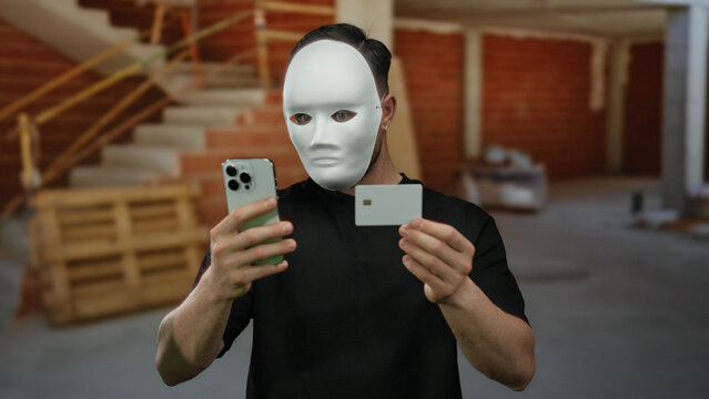 Young man with beard in mask holding smartphone and card at indoor construction site suggesting identity concealment and financial transaction.