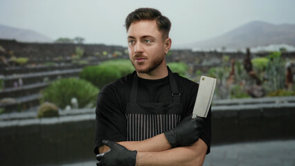 Young man with beard in apron and gloves holding butcher knife standing in outdoor park setting, showcasing culinary skills and preparing for cooking demonstration.