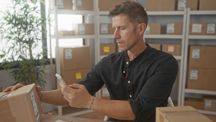Obraz premium Man holding cardboard parcel and smartphone with bare hands while scanning label in a building; efficiency focus.