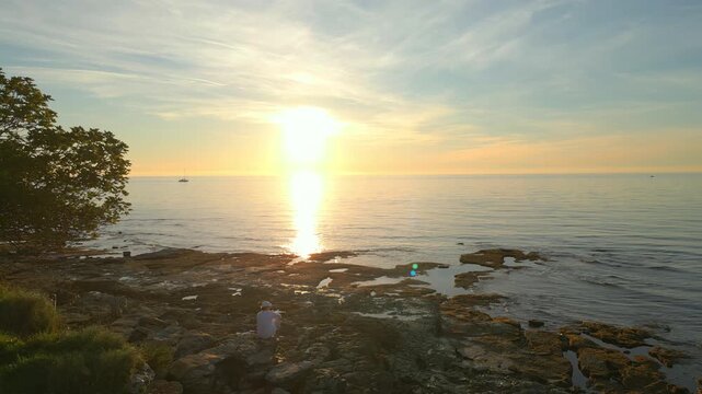 A person sits on rocky shores at sunset, gazing at the horizon. The serene ocean and golden sky evoke mindfulness and reflection, offering a peaceful escape from daily life.