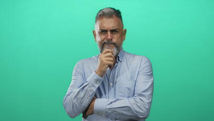 Man with hand on chin and crossed arms in mint green studio wearing a light blue shirt and gray beard visible, head tilted slightly in thinking pose; thoughtful reflection.