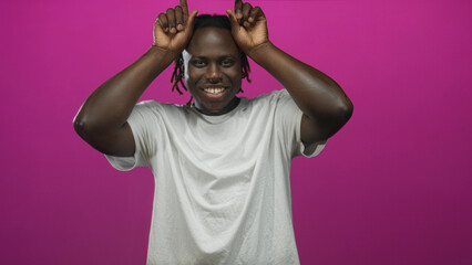Young african american man with hands making horn gesture in studio pink backdrop wearing white t shirt and dreadlocks; playful mischief confidence.
