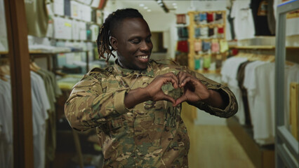 Man soldier wearing camouflage uniform and dog tags forms a heart with hands while smiling in a retail clothing store building lined with racks; pride duty.