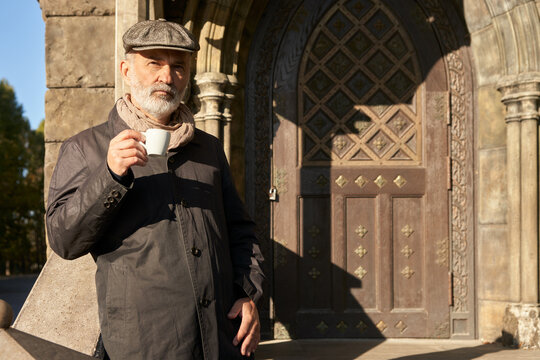 An elegant grey-bearded senior citizen with a cup of morning coffee at the entrance to an ancient castle.