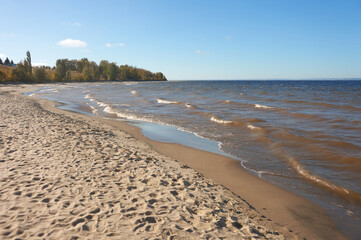 The sandy shore of a wide river with waves crashing onto the beach. Numerous shoe prints are visible in the sand. Autumn. Background image.