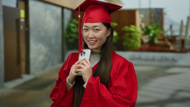 Young woman in red graduation gown smiling and holding smartphone on urban street backdrop with greenery