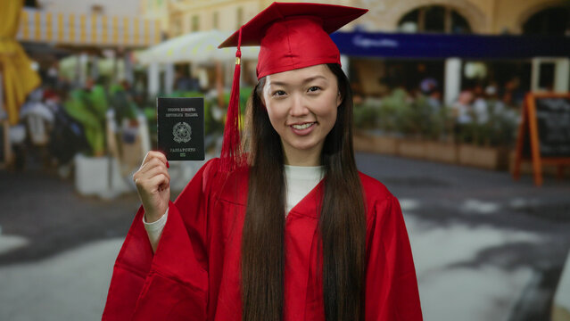 Fototapeta Woman in red graduation gown holding italian passport on a bustling city street cafe terrace showcasing cross-cultural achievement