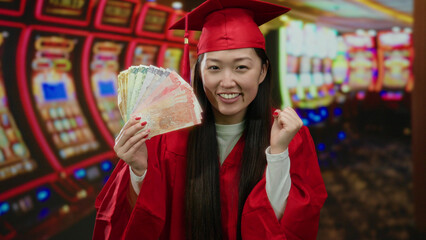 Woman in red graduation gown holding philippine banknotes in a casino with slot machines in the background, smiling and celebrating indoors.